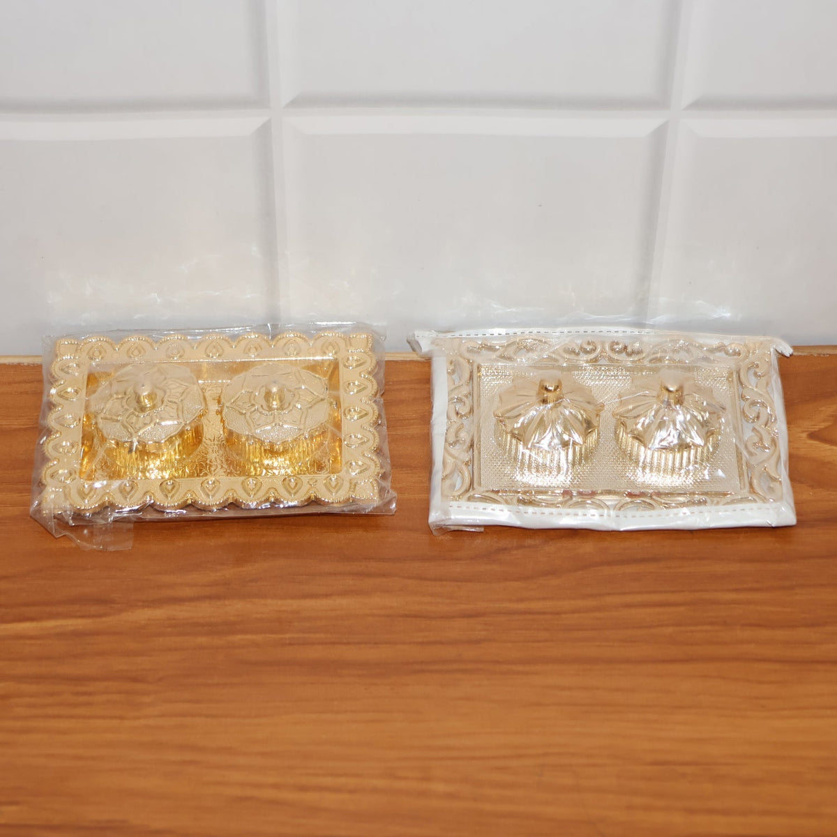 Two ornate rectangular pooja thalis, one gold and one silver, both in clear plastic packaging, on a wooden surface.