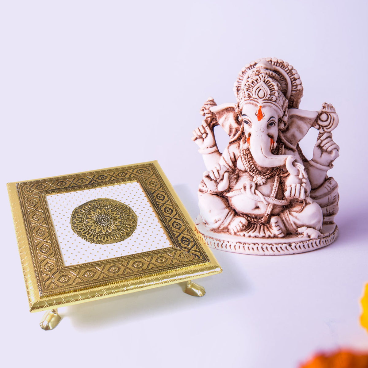 A golden square pooja chowki next to a white Ganesha idol, with a hint of yellow flower petals in the foreground.