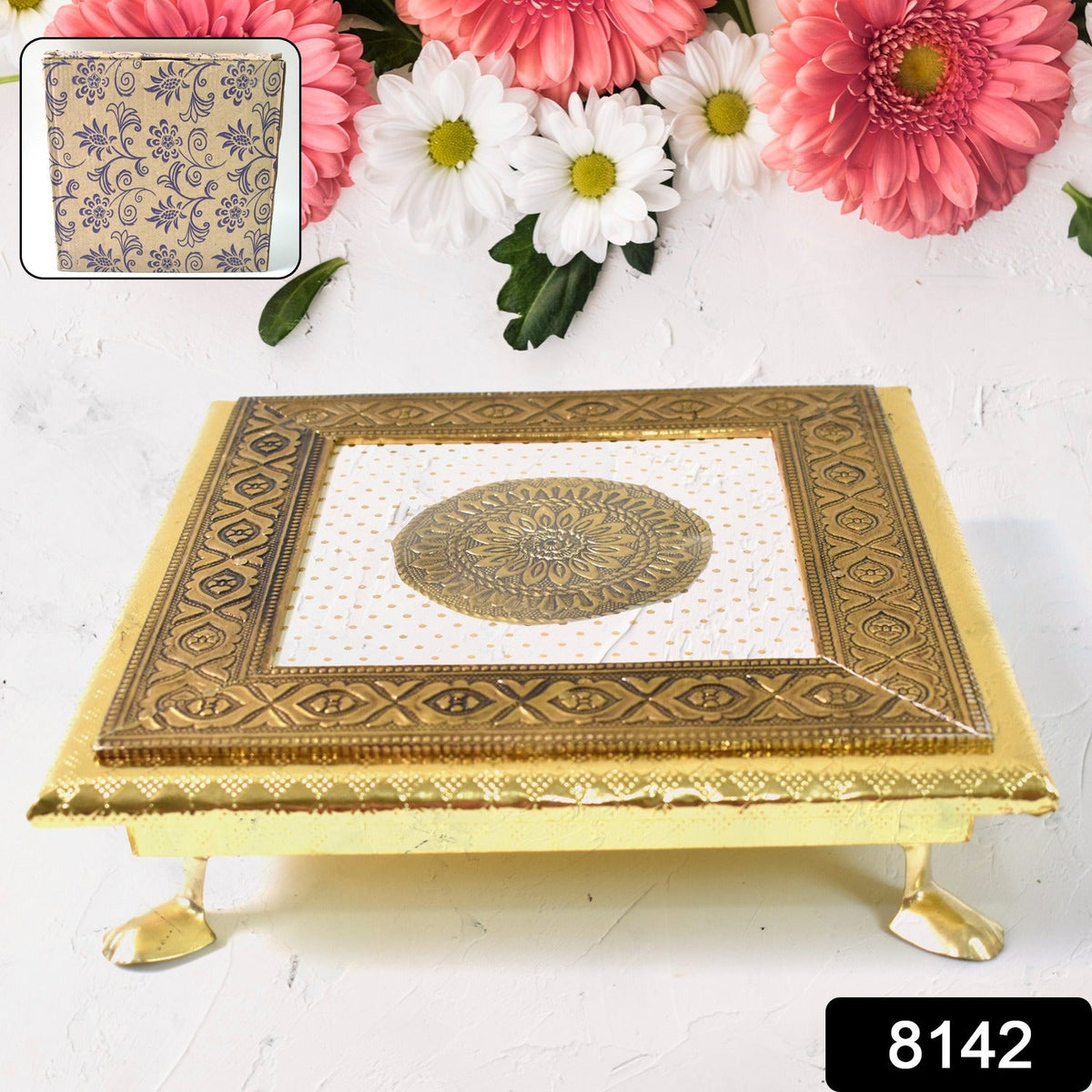 A golden square pooja chowki with an intricate mandala design, against a background of pink and white flowers. An inset