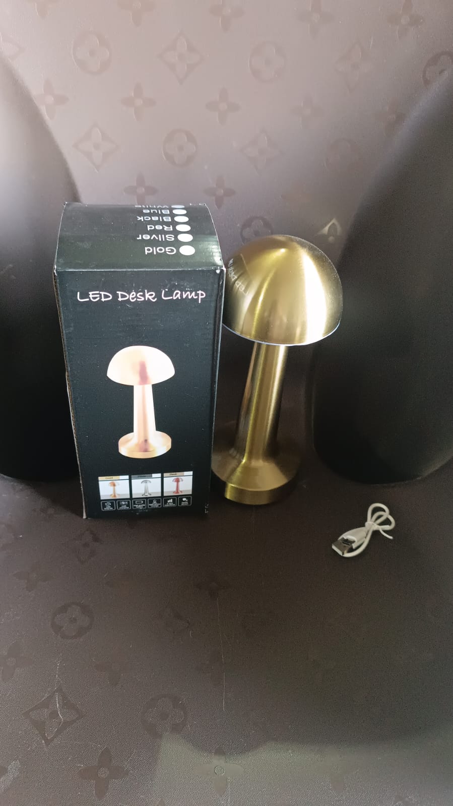 A black box for an LED desk lamp, a gold mushroom lamp, and a white charging cable are on a dark patterned surface.
