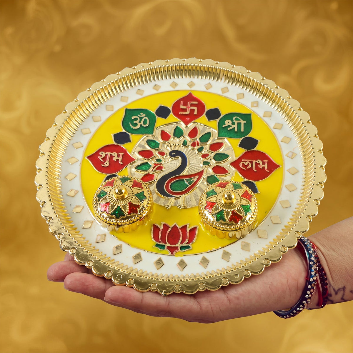 A person's hand holding a golden pooja thali with a peacock design and two small bowls.