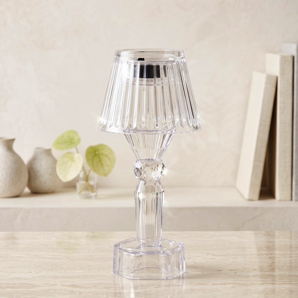 A single clear acrylic miniature table lamp with a ribbed shade on a marble surface, with decorative vases and books in the