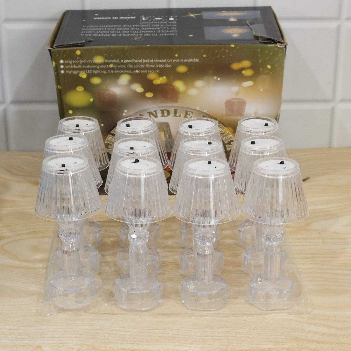 Ten clear acrylic miniature table lamps in plastic packaging, arranged in front of a brown cardboard box with product