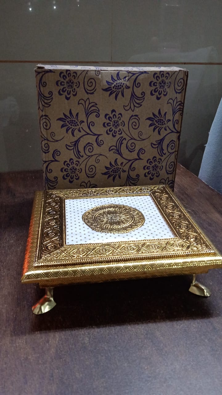 A golden square pooja chowki with a patterned brown box standing upright behind it, all on a wooden surface.